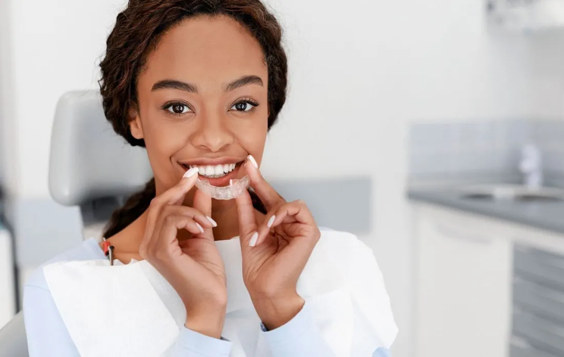 Patient smiling while holding a clear Invisalign aligner in the dental chair